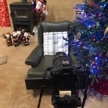 a digital slr camera on a tripod beside a christmas tree, aimed at a green chair with small santa figures on the floor in front of a woodstove. stockings hanging from a stand
