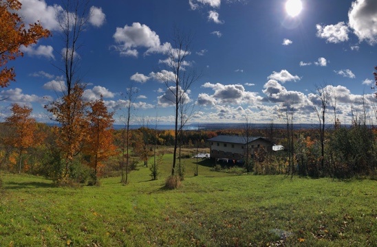 fluffy clouds in a blue sky with the sun shining. green grass on a slope with golden leaves on the trees. a house is below and the sun is reflecting off part of the black roof. there is water and hills in the distance