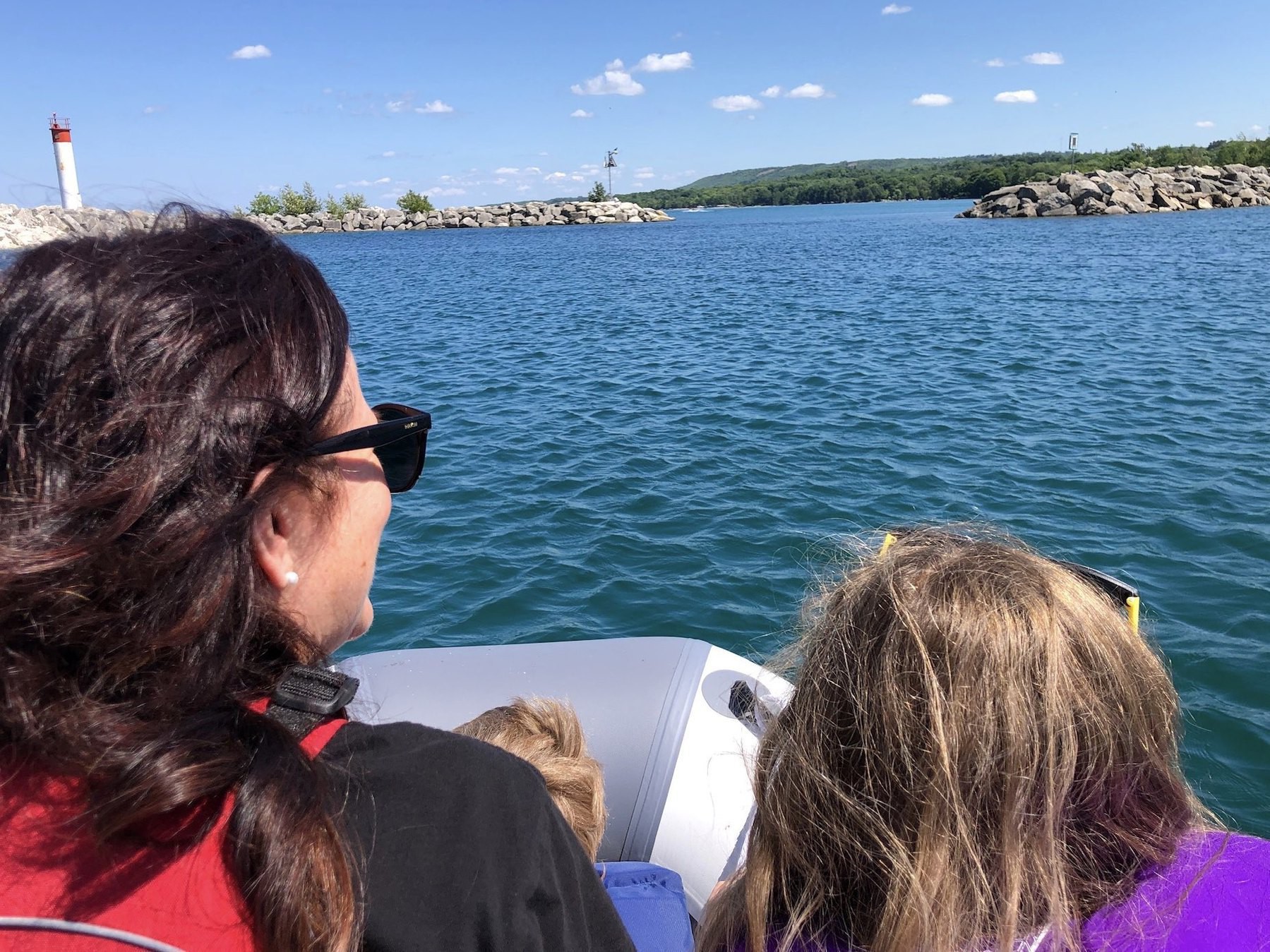 a woman and her two grandchildren wear lifejackets while riding in a small zodiak. Large rocks make a breakwall and trees are visible in the distance