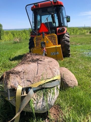 large rock with 2 yellow straps around it sitting behind a tractor with a winch