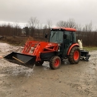 orange tractor with cab, front end loader and a snowblower on the back. it is sitting on a wet, muddy driveway with some tools sticking out of the bucket