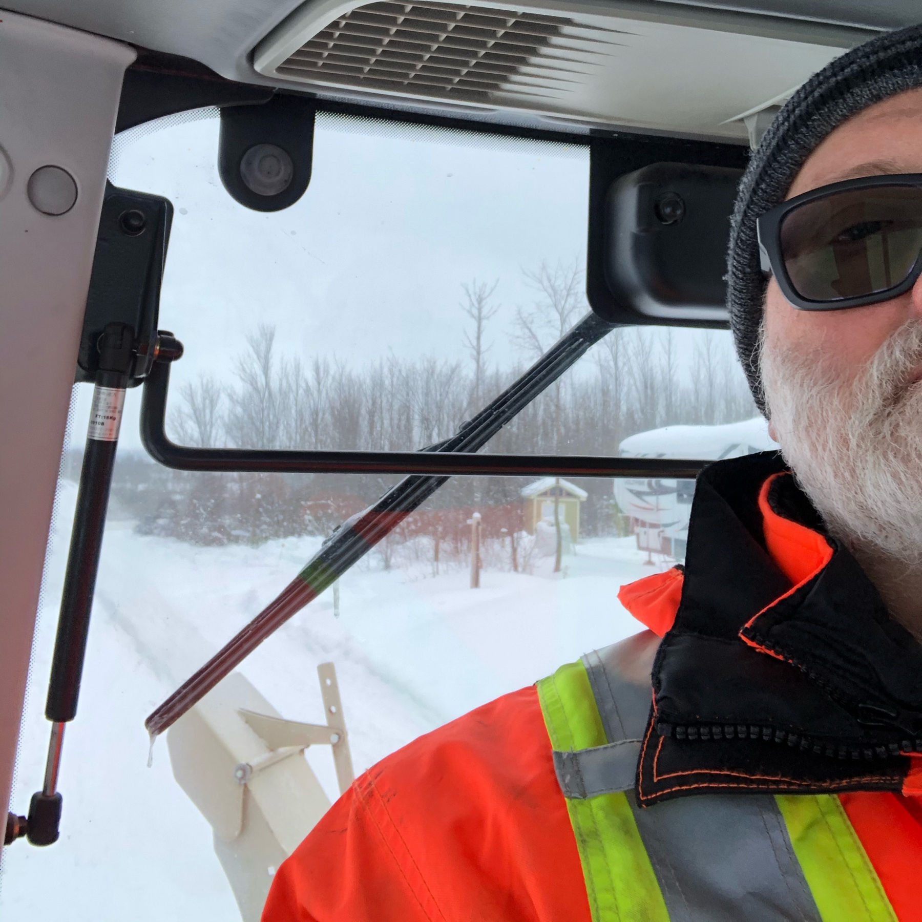 man with beard, wearing a hi vis jacket. he is sitting in the cab of a tractor. snow is coming out of the chute behind the tractor. 