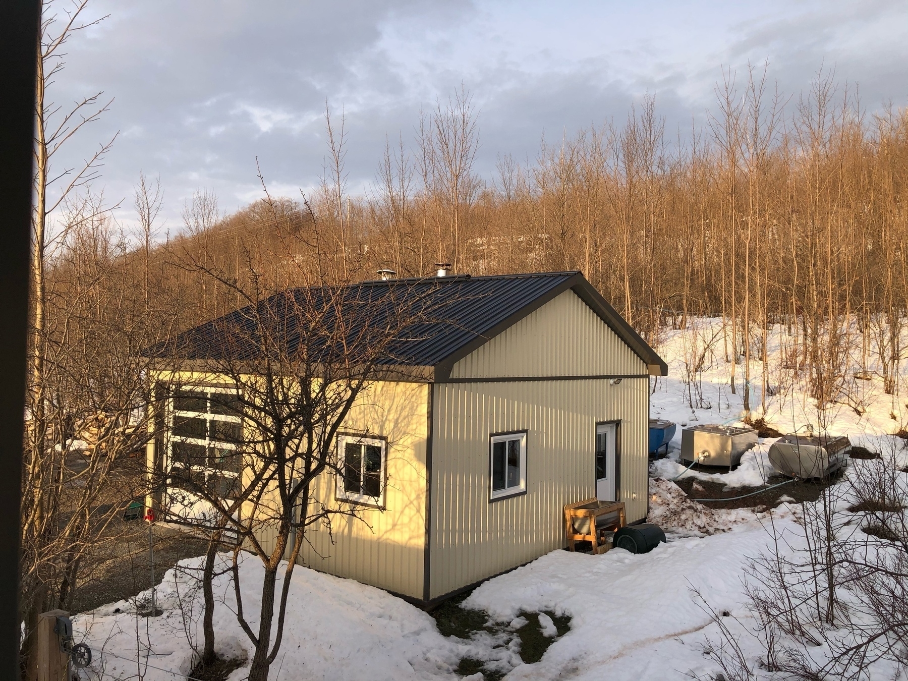 small building surrounded by snow and bare ground. there are stainless steel tanks behind. the hillsode behind is covered in trees