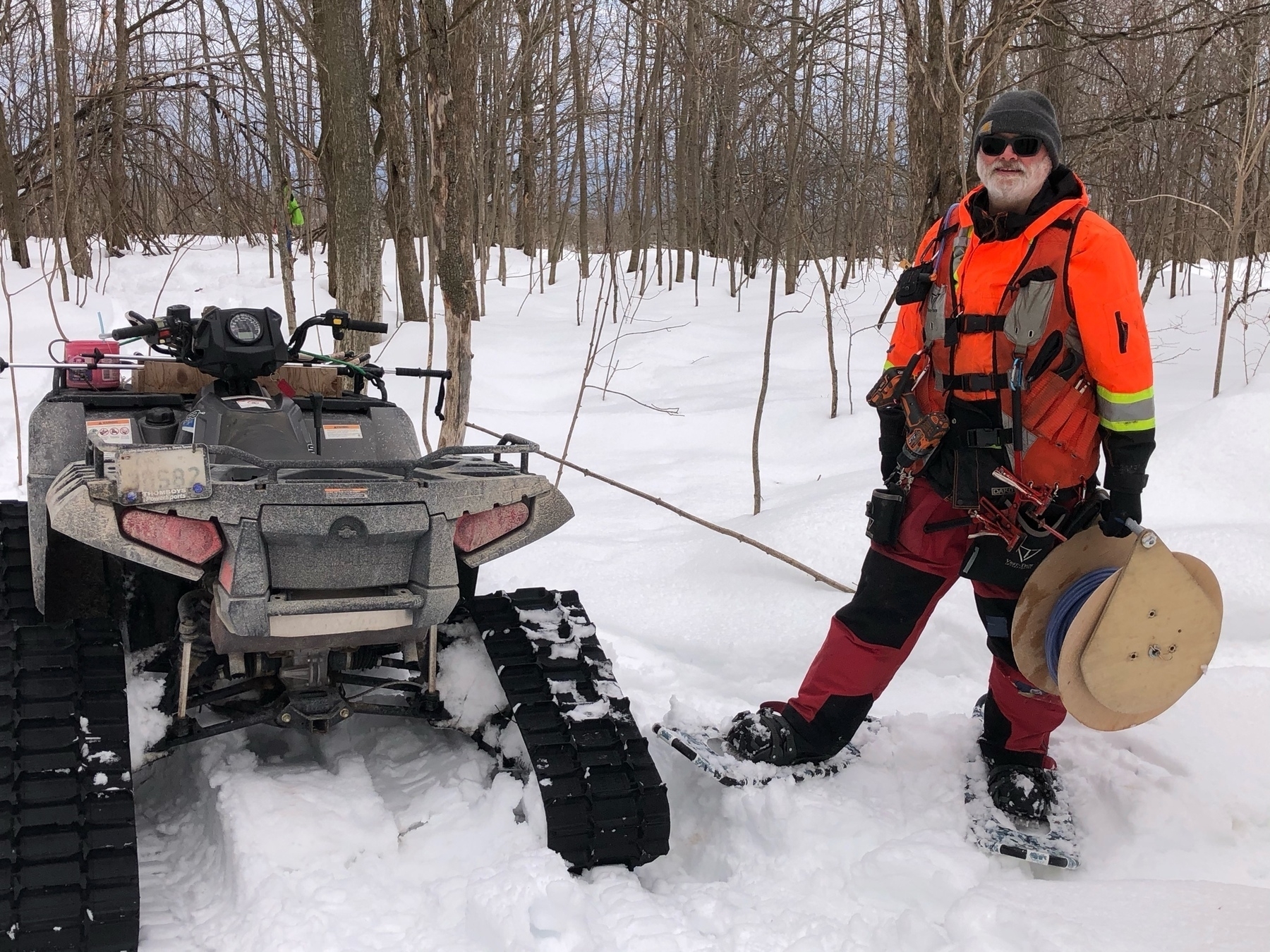 bearded man in fluorescent vest, red pants and snowshoes. he is wearing a toque and gloves and standing beside an atv with tracks. the ground is snow covered and he is surrounded by small trees 