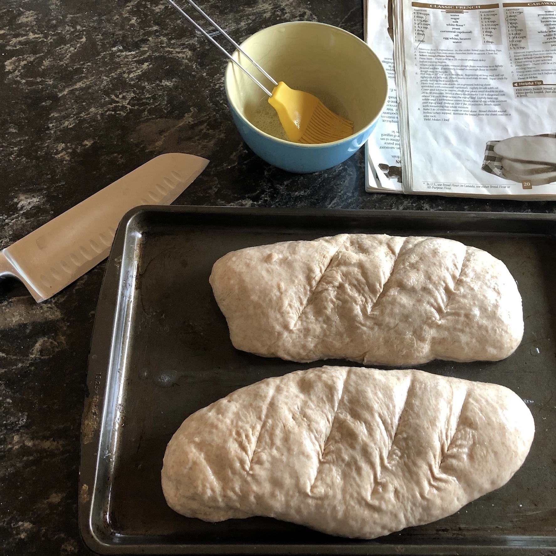Two dough rolls, scored diagonally on top with a blue dish of egg wash and yellow brush beside them