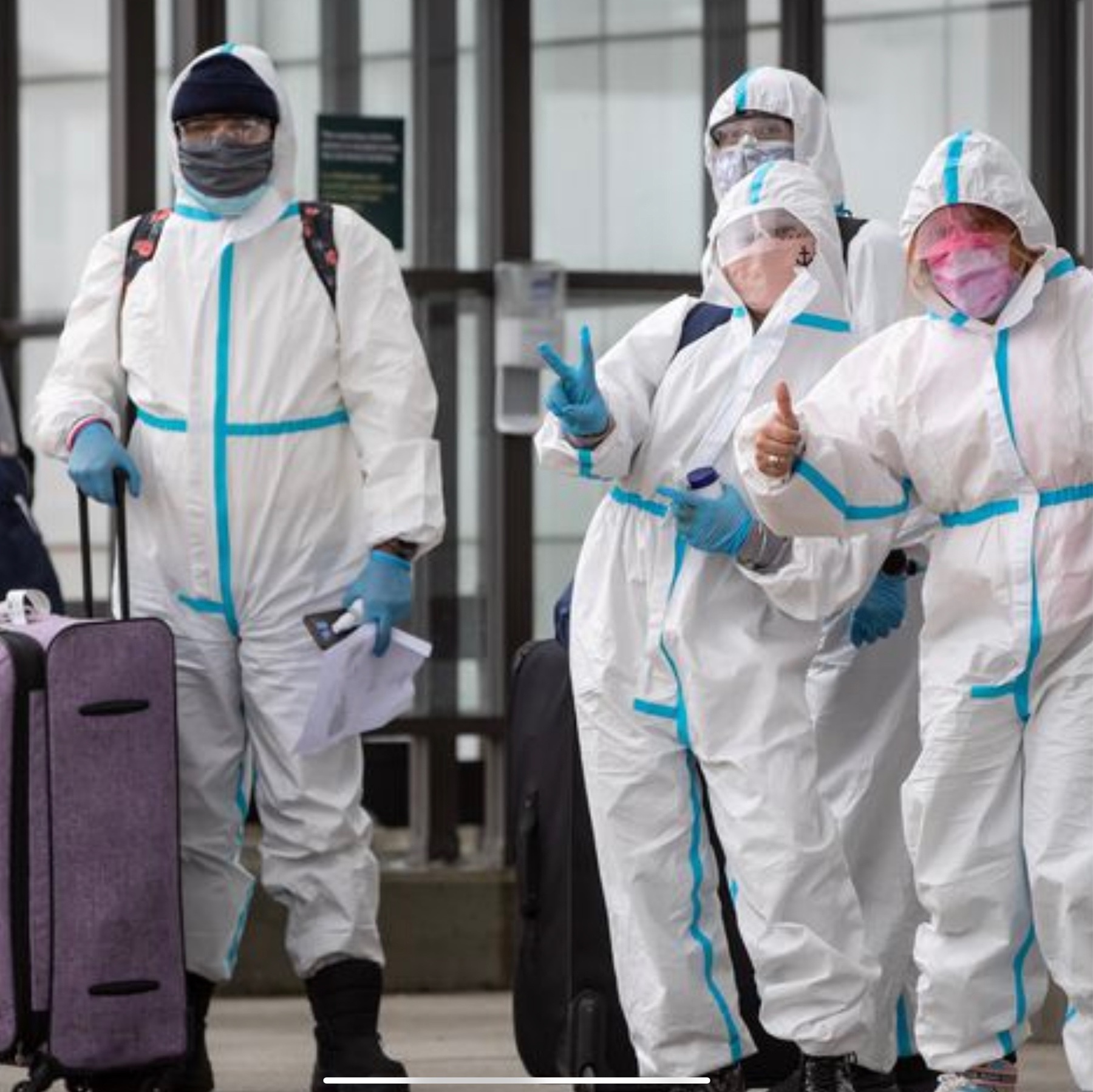 4 people standing outside an airport giving thumbs up to the camera. They are dressed head to toe in white coveralls, and wearing safety glasses and face masks