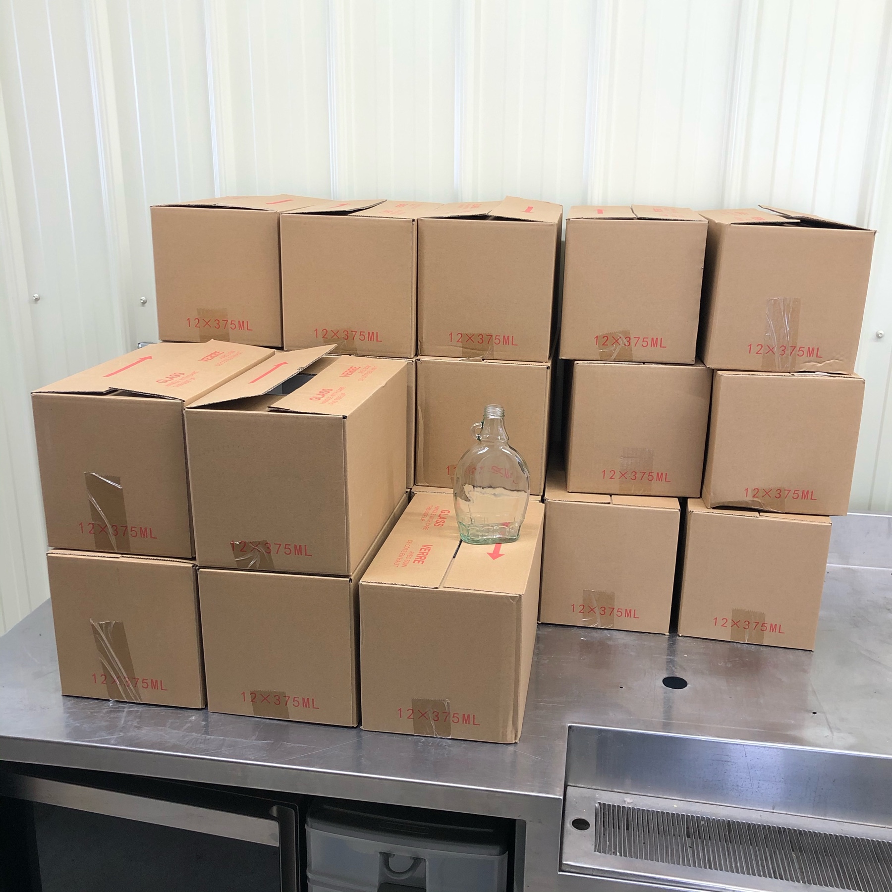 20 plain, cardboard boxes piled on a stainless steel counter. there is one bottle on display. 