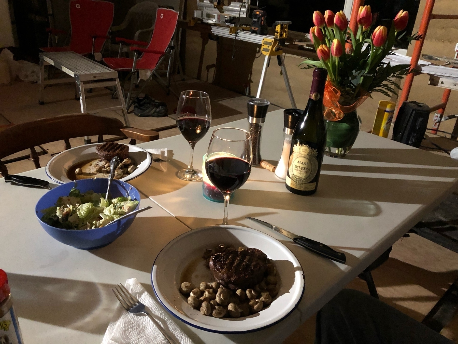 steak and mushrooms on 2 plates sitting on a wooden table. there are orange tulips in a vase on the table