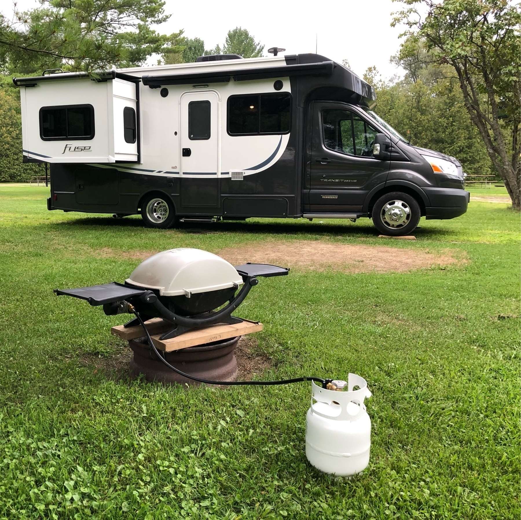 small motorhome with a bbq in front. a pine tree is overhead 