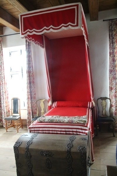 red, canopy-covered bed with a steamer trunk at its foot. pink and white curtains hang on the tall windows