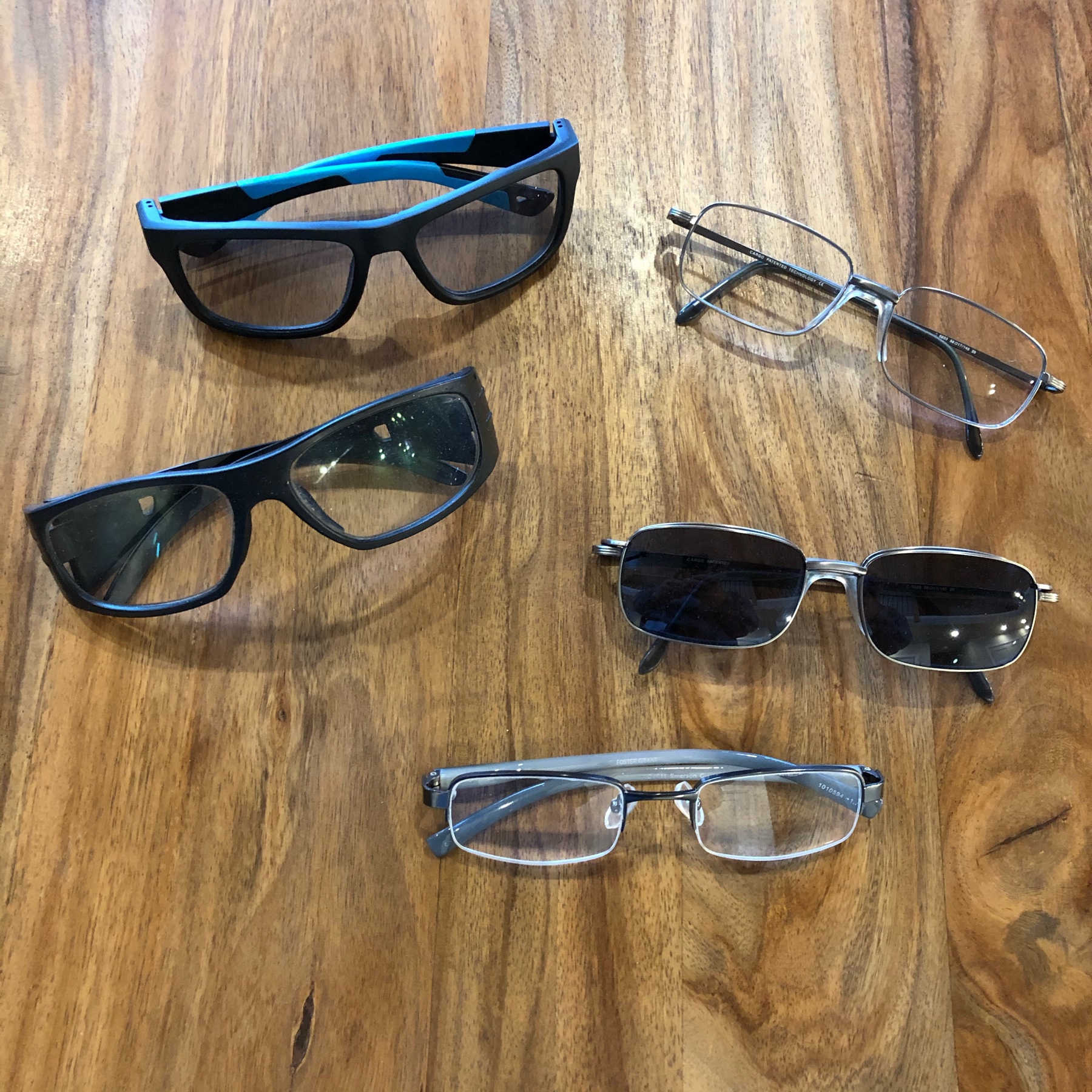 Five pair of glasses. One pair is sunglasses and two pair are safety glasses. they are sitting on a wooden table