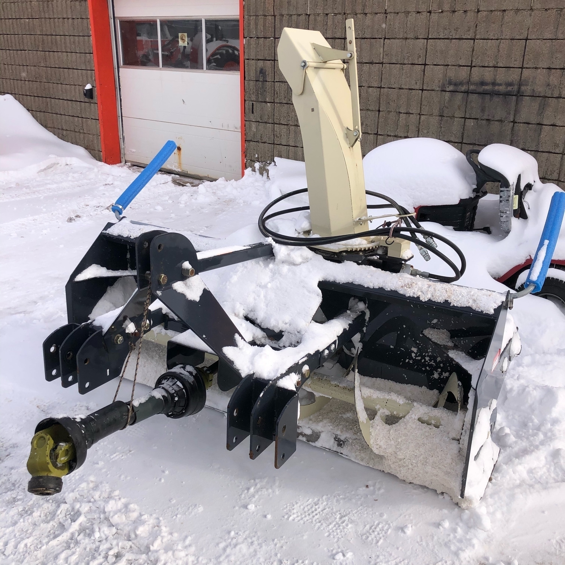 a snow-covered snowblower  for a tractor sits in front of a building