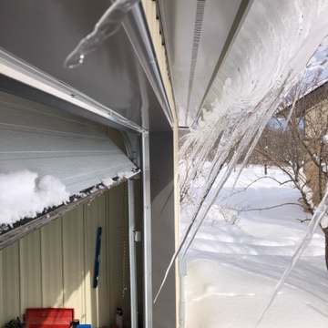 icicles hanging at about 30 degrees from vertical. other snow and ice is curling off the edge of a roof on a building with dark grey trim. there is a fruit tree in the background and snow covering the ground