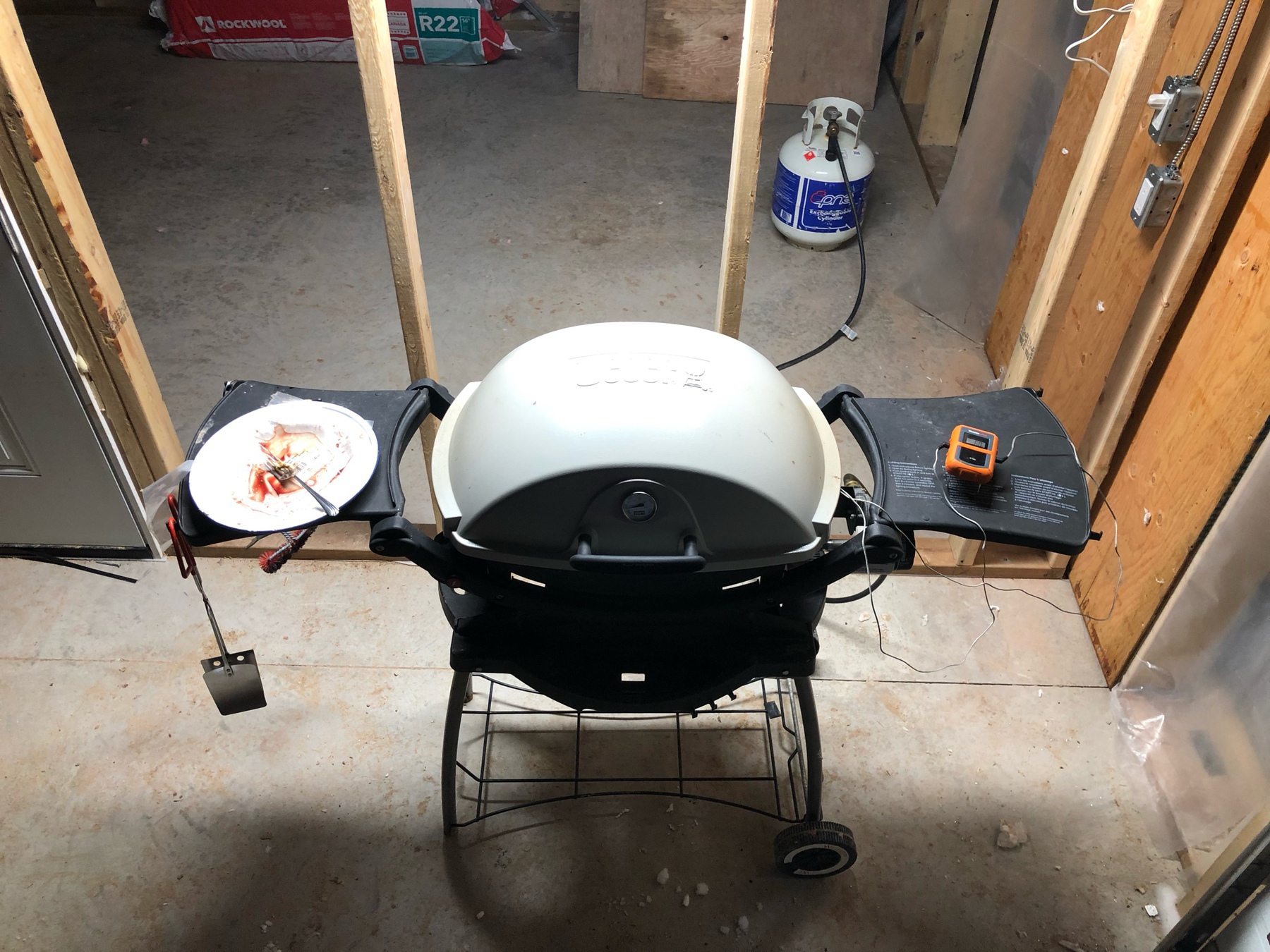 a small bbq in front of a stud wall. there is a propane tank in the distance